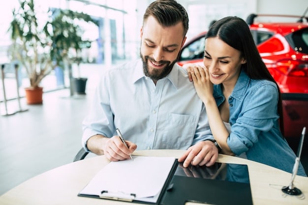 A close-up of a contract related to car financing being signed by a man while a woman looks on, both smiling. A car key and a pen are on the table.