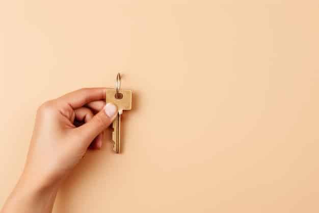 Close-up shot of a person's hand holding a key to a new house. The background is slightly blurred, showing a smiling couple walking into the house. The key has a small tag attached with the words 